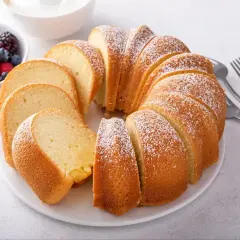 Pyszna, wilgotna babka z cukrem pudrem, pokrojona w plastry, obok miseczki z owocami leśnymi i śmietaną.