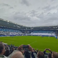 Trybuny stadionu wypełnione kibicami Karlsruher SC. Fani tworzą ogromną oprawę z napisami i sylwetką zawodnika.