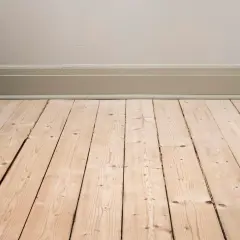 Ett ljust, lutat furugolv med synliga ådringar och kvistar. Golvet möter en ljusbeige vägg med en gråmålad golvlist.