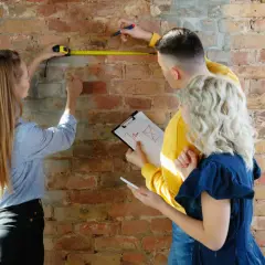 Para planować kredyt na remont mieszkania, trójka osób mierzy ścianę i notuje wymiary.