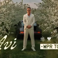 Mężczyzna w garniturze stoi przed czarnym samochodem wśród kwitnących drzew. Napis "Avi" i "WPR TOUR".