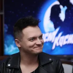 Uśmiechnięty mężczyzna w skórzanej kurtce, na ekranie za nim widać napis "Nocny Kochanek" i sylwetkę pary.
