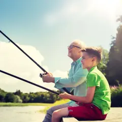 Abuelo y nieto disfrutan de un día de miños pesca, con cañas en mano y sonrisas en el rostro, bajo un cielo azul y soleado.