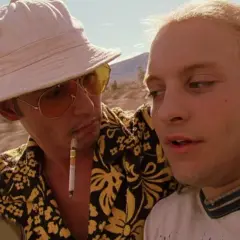 Dwóch mężczyzn w samochodzie, jeden z papierosem w ustach, w stylu filmów o uzależnieniach. Pustynny krajobraz w tle.