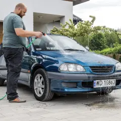 Mężczyzna myje niebieski samochód na posesji, używając węża ogrodowego. Woda spływa po karoserii i tworzy kałuże na podjeździe.
