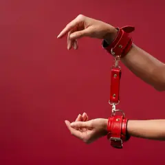 Czerwone kajdanki na nadgarstkach, symbolizujące intymną relację BDSM, na tle bordowej ściany.