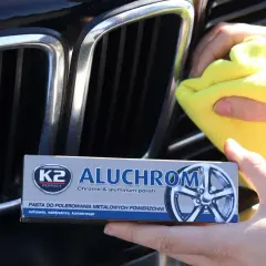 Ręce polerują chromowaną atrapę chłodnicy samochodu żółtą ściereczką. Obok widać opakowanie pasty K2 Aluchrom.