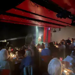 En föreställning på soppteatern Stockholm. En ensam artist står på scenen i strålkastarljus, publiken sitter vid dukade bord.