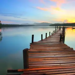 A wooden pier stretches into a calm lake at sunset, showcasing great depth of field photography. The sky transitions from blue to vibrant orange, reflecting on the water.