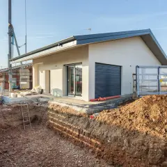 Budowa domu: wykop pod fundamenty, koparka i częściowo wykończony budynek.