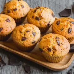 Sei muffin allo yogurt con gocce di cioccolato su un vassoio di legno, circondati da pezzi di cioccolato fondente.