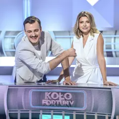 Uśmiechnięty mężczyzna i kobieta w białej sukience, którzy prowadzą Koło Fortuny, stoją przy pulpicie.