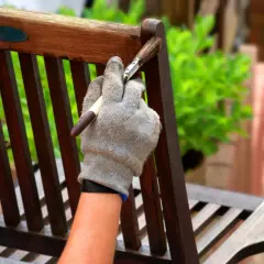 Ręka w rękawicy nakłada lakierobejcę do mebli ogrodowych na drewnianą ławkę, odnawiając jej wygląd.