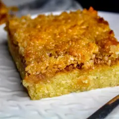 En bit saftig toscakaka i långpanna med karamelliserad mandeltopping. Serveras på en vit tallrik med en sked.