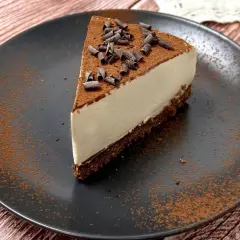 Una fetta di cheesecake al caffè, cosparsa di cacao e riccioli di cioccolato, su un piatto nero.