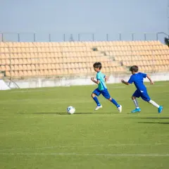 Dwóch chłopców w strojach piłkarskich biegnie po zielonej murawie stadionu, ćwicząc jedną z popularnych dyscyplin sportowych po angielsku.