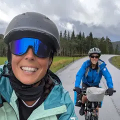 Dos ciclistas disfrutan de la ruta EuroVelo 6, con un paisaje verde y montañoso bajo un cielo nublado.