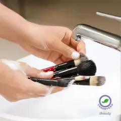 Mani che lavano pennelli da trucco insaponati sotto un rubinetto cromato. Un gesto di cura per un make-up impeccabile.