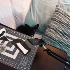 A small black dog watches as someone uses adaptive sewing tools, including a rotary cutter and pins, on a cutting mat.