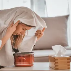Kobieta wykonująca inhalację parową nad garnkiem z wodą, przykryta ręcznikiem.