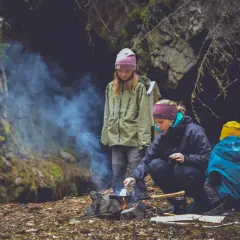 Familj lagar mat ute vid en lägereld i skogen. Rök stiger från elden där en kvinna steker något.