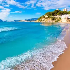 Una playa de arena dorada y aguas turquesas, con edificios blancos y vegetación en la ladera, que evoca las 10 mejores playas de Alicante.