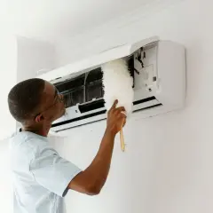 Un homme nettoie son climatiseur avec un plumeau. Un entretien régulier est essentiel pour savoir entretien climatisation tous les combien.