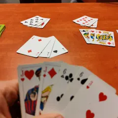 Gracz trzyma karty do gry w remika: walet serce, walet karo, szóstka trefl, szóstka pik, trójka serce. Na stole leżą inne karty.