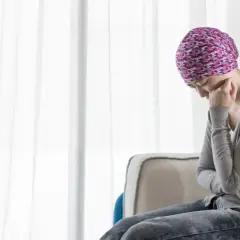 Kobieta w chuście na głowie siedzi przy oknie, zamyślona, być może doświadczając skutków ubocznych chemioterapii.
