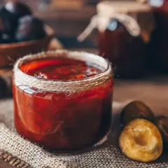 Ein Glas Pflaumenmus, wie Oma es einkochte, steht auf rustikalem Leinen. Frische Pflaumen liegen daneben.