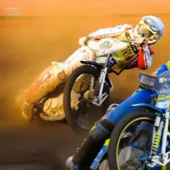Dwóch zawodników ściga się na torze, ich motocykle żużlowe unoszą kurz.
