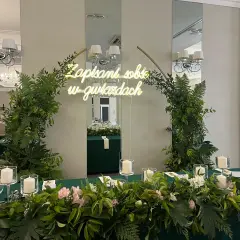 Dekoracja ścianki za parą młodą z neonem "Zapisani sobie w gwiazdach", otoczona bujną zielenią i świecami.
