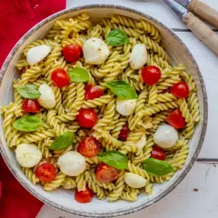 Un piatto invitante di pasta fredda al pesto, arricchito da ciliegini succosi e mozzarelline fresche, guarnito con basilico profumato.