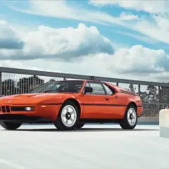 Pomarańczowe BMW E26, ikona lat 80., prezentuje się na tle błękitnego nieba z chmurami.