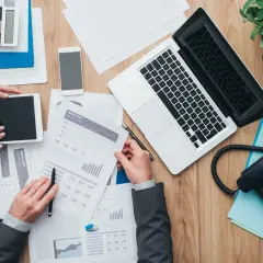 Widok z góry na biurko z laptopem, telefonem, kalkulatorem, tabletami i dokumentami finansowymi.