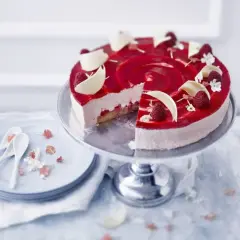 En vacker hallon cheesecake med hallonspegel, vita chokladdekorationer och färska hallon. Serveras på ett tårtfat med en skiva borttagen.