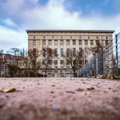 L'imponente edificio del Berghain, con la sua facciata austera e le finestre scure, si staglia sotto un cielo nuvoloso. Un cancello metallico e alberi spogli circondano l'area, suggerendo un percorso misterioso per chi cerca come entrare al Berghain.