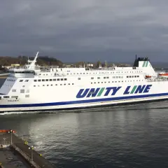 Biały prom Unity Line "Polonia" płynie po wodzie, w tle widać port i zachmurzone niebo.