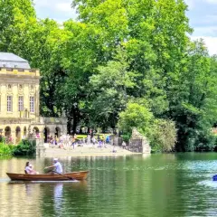 Ein sonniger Tag am Monrepos See. Menschen rudern in einem Boot, während im Hintergrund ein prächtiges Gebäude und üppige Bäume zu sehen sind.