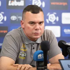 Trener Jagiellonii w szarej koszulce z logo klubu, siedzi przy mikrofonach podczas konferencji prasowej.