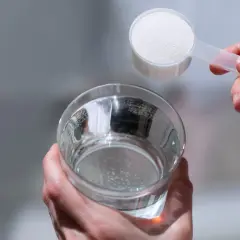 Dłoń wsypuje miarkę hydrolizowanego kolagenu do szklanki z wodą, przygotowując zdrowy napój.