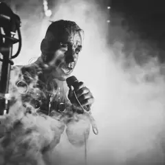 Lindemann w dymie, z mikrofonem w dłoni, w mrocznym klimacie. Jego twarz jest intensywna, jakby śpiewał "Knebel".