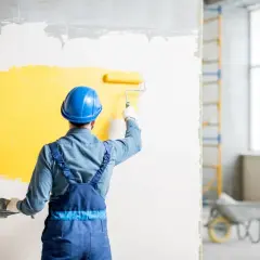 Malarz w niebieskim kasku maluje ścianę na żółto wałkiem.