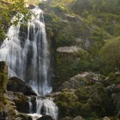 La majestuosa cascada de Belelle cae en cascada por rocas cubiertas de musgo, con gente disfrutando del paisaje y el agua.
