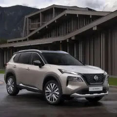 Elegancki, beżowy Nissan X-Trail 2015 zaparkowany przed nowoczesnym budynkiem w górach.