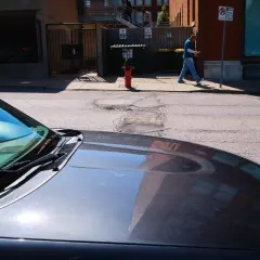 A man walks past a fire hydrant on a sunny day, his reflection visible in the hood of a car. 40mm street photography captures the urban scene.