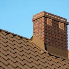 Ceglany komin do kominka na brązowym dachu pod błękitnym niebem.