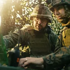 Dwóch żołnierzy w mundurach analizuje dane na laptopie. W tle słońce przebija się przez liście. Skróty wojskowe polskie są kluczowe w ich pracy.