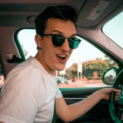 Joven con gafas de sol sonríe emocionado al volante de su coche novel, listo para una aventura.