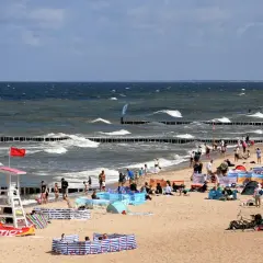 Plaża nad Bałtykiem pełna ludzi, fal i parawanów. Widać ratownika, sprzęt wodny i tablicę informacyjną. Ceny nad morzem pewnie wysokie, ale widok wart zachodu.